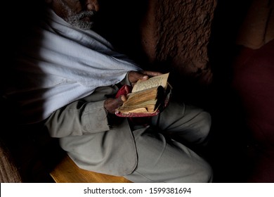 Lalibela, Ethiopia, Africa - November 21 2019: Priest Wearing Old Tatty Grey Suit With Frayed Cuffs Seated In Partial Shade In A Church Doorway Reading A Bible Script Book In Lalibela,Ethiopia, Africa