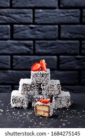 Lamingtons Vanilla Sponge Mini Cakes With Strawberry Jam Layer Inside, Coated With Chocolate And Coconut On Top On A Dark Concrete Table In Front Of A Black Brick Wall, Portrait, Copy Space