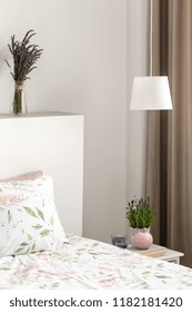 Lamp Above Cabinet With Plant Next To Bed With Sheets In Bedroom Interior With Lavender Flowers. Real Photo