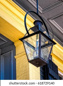 Lamp
A Black Lamp Hangs From The Ceiling Of A Yellow Painted  Porch In New Orleans. It Has A Glass Chimney And Uses Gas To Make The Light. The Chimney Is Surrounded By Four Glass Panes. 