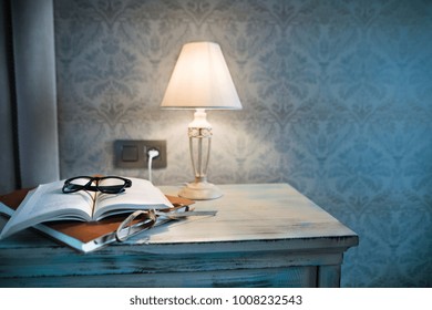 A Lamp And A Book On A Bedside Table In A Hotel Room.