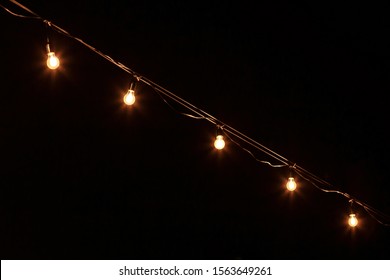 Lamp Garlands On A Black Background Outdoor. Hanging Light Bulbs Backdrop. Lighting Decor. Retro And Vintage Concept. Copy Space.