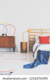 Lamp On Wooden Stool Next To Bed With Red Cushion And Blue Blanket In Bedroom Interior. Real Photo