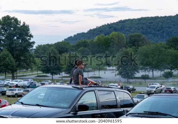 Lancaster, Pennsylvania - July 3, 2021: A lot of\
cars parked on a hillside and some kids sitting on a car roof\
waiting for the fireworks show to\
start.