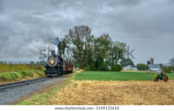 Lancaster, Pennsylvania, October 2019 -\
Norfolk and Western Steam Locomotive no. 382 Freight and Passenger\
Train Blowing Smoke and Steam as it Passes a Period Farm Tractor in\
the Countryside