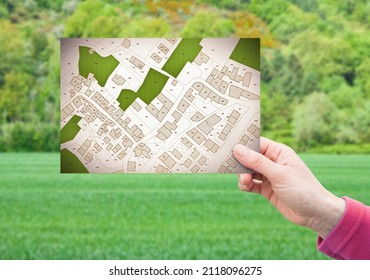 Land plot management - real estate concept with a vacant land on a green residential area available for building construction with hand holding a postcard about an imaginary cadastral map