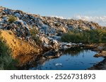 The landfill displays large mounds of mixed waste surrounded by patches of greenery and a winding stream, reflecting the environmental impact of waste disposal