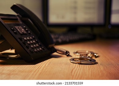 Landline Phone And Handcuffs On The Table, Close-up. Office Desk In A Dark Night Room With Computer Monitors