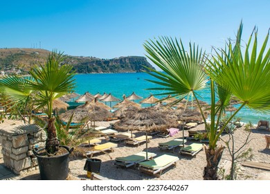 Landscape of beautiful clean sand and pebble beach with beach umbrellas and sun loungers. Green palm leaves in the foreground and  mountains at background. Himare. Albania. Ionian Sea. Sunny landscape