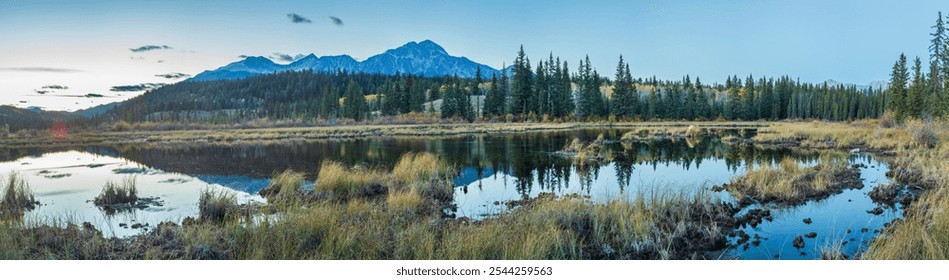 A landscape in Canada showing a clear lake reflecting dense evergreen forests and majestic mountains under a pastel sky. The calm water conveys the peaceful atmosphere of this untouched wilderness. - Powered by Shutterstock