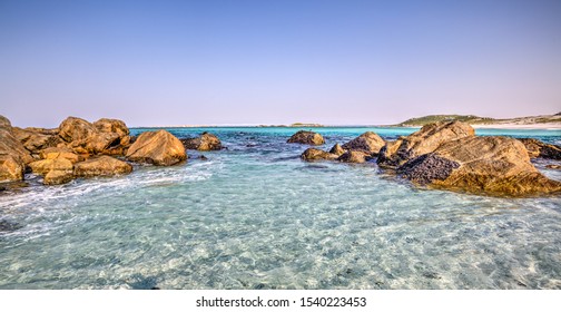A landscape of the clear ocean which looks like tropical waters. Rocks on the sides, beach and islands in the background.