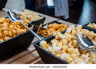 Landscape Close Up Photo Of Many Fancy Bowls Of Pop Corn With Different Flavors (butter, Salt, Bbq) With Spoons Laying On The Pop Corn On A Wooden Table At A Crowded Event.