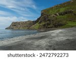 A landscape coastal image of Talisker Bay Beach on the Isle of Skye, Scotland. The black and silver stretch to steep rocky cliffs over which a waterfall flows, all beneath a blue sky.