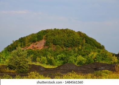 
The Landscape Consists Of A Burnt Mine Waste Heap, Sheltered By Green Vegetation And Heaps Of Coal Sludge.