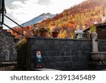 Landscape of decorative garden gnome dwarf with hat on the windowsill of a house on the green lawn among the Italian Alps Mountains in autumn with trees yellow and orange leaves in the background