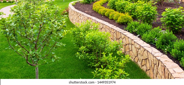 Landscape Design In Home Garden, Beautiful Natural Landscaping With Flower Beds And Lawn. Panoramic View Of Green Landscaped Yard. Scenery Of Trees, Plants And Stones In Residential House Backyard.