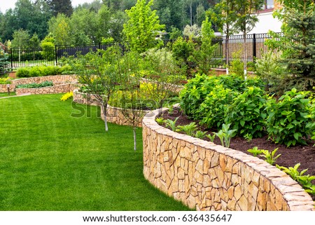 Landscape design in home garden,  landscaping with plants, trees and stones in residential house backyard. Beautiful landscaped back yard with retaining walls and green lawn in summer. Nature theme.