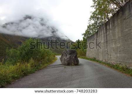 Landscape image of a large boulder blocking a narrow asphalt road surrounded by lush greenery and mist-covered mountains, captured in Norway