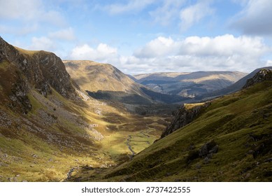 A landscape image looking down into a deep valley from the top of Corrie Fee in the Scottish Highlands. Small white clouds in the blue sky cast shadows along the green valley floor as the Scottish Highlands rise high in the distance.