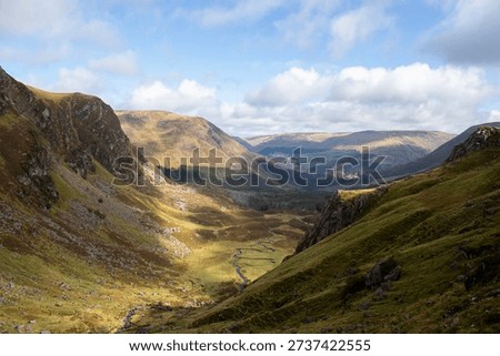 A landscape image looking down into a deep valley from the top of Corrie Fee in the Scottish Highlands. Small white clouds in the blue sky cast shadows along the green valley floor as the Scottish Highlands rise high in the distance.