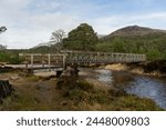 A landscape image of a narrow wooden bridge spanning Lui Water near Derry Lodge in the Scottish Highlands.  Derry Cairngorm rises above the green pine trees in the distance towards a blue sky.