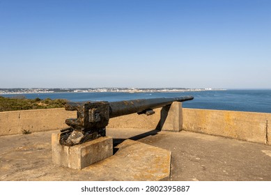 A landscape image of a second world war German gun, at Noirmont point, Jersey Channel Islands, The barrel of the gun is 8 meters, and is on top of a bunker. Behind and in the distance is St Aubins bay and the town of St Helier. The sea is calm and blue, the sky is clear blue.