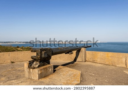 A landscape image of a second world war German gun, at Noirmont point, Jersey Channel Islands, The barrel of the gun is 8 meters, and is on top of a bunker. Behind and in the distance is St Aubins bay and the town of St Helier. The sea is calm and blue, the sky is clear blue.