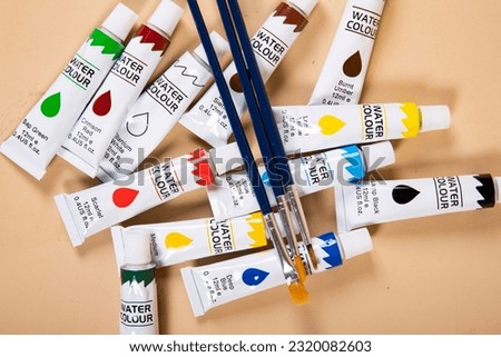 A landscape image of watercolour paint tubes sprawled all over a beige background, with three fine tip paint brushes sitting on top.