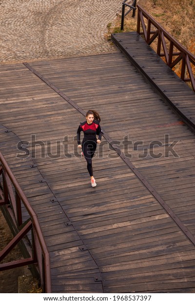 landscape, one young woman running, jogging\
outdoors over a wood\
bridge.