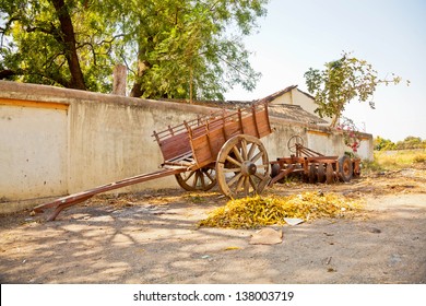 Landscape at the outskirts of a Gujarat village in India of a wall lined farmhouse with farm machinery and parked bullock cart under the shade of trees and a pile of autumn leaves