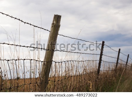 A landscape photograph of a barb wire fence leading down the horizon of an open field. A blue sky with white clouds fills the sky.