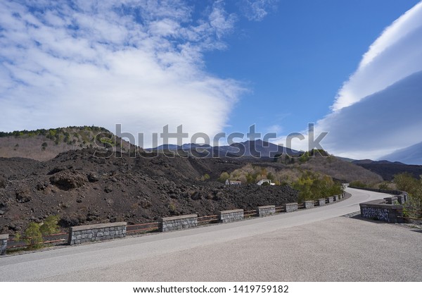 Landscape picture on road to mount etna, biggest active\
european volcano in italian island sicily in the summer sunny but\
cloudy day. Asphalt road in black lava rock field from last volcano\
eruption. 