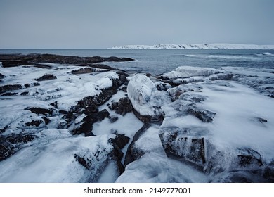 Landscape with rocky shore on the ice-free northern Arctic Ocean in deep twilight. Frozen stones and ice in the foreground in cloudy weather.