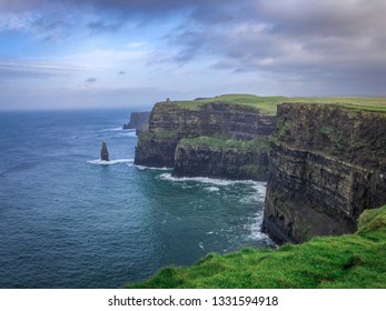 Landscape Shot Of The Cliffs Of Moher In Ireland - On A Cold Winter Day With Green Grass, Cragged Rocks, Bright Blue Water And Cloudy Skies Overhead
