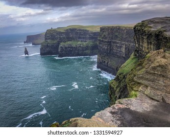 Landscape Shot Of The Cliffs Of Moher In Ireland - On A Cold Winter Day With Green Grass, Cragged Rocks In The Foreground, Bright Blue Water And Cloudy Skies Overhead