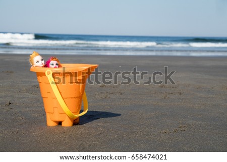 Landscape shot of two barbies in an orange toy bucket with the waves as background