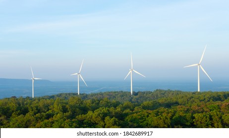 Landscape Sun Set Shot Of Wind Turbines Farm On The Green Grass Field With Clear Blue Sky And Mountain At The Background. Alternative Green Energy Which Generate Electricity From Natural Wind Power.