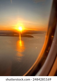 A landscape of the sun setting over remote islands in the ocean through the view of an airplane window. The gold sun rays reach across the sky and water, reflecting off of the sparkling ocean.