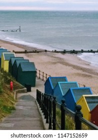 Landscape view down steep pathway to beach with bright coloured wood beach huts along promenade in front of beautiful sandy beach with sea waves and railing walking from cliffs on summer holiday