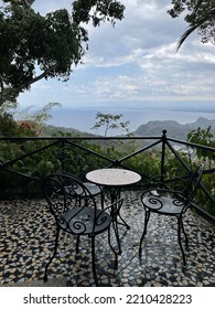 A Landscape View Of The Ocean In The Distance From A Small Patio That Has Chairs And A Table Set Back In The Jungle