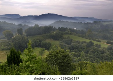 Landscape view of a village in the countryside of Spain. Green trees over hills and foggy mountains in the distance. Bright sun light shines behind clouds creating a soft purple and blue sky at dusk. - Powered by Shutterstock