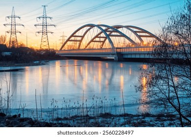 Landscape with a winter frozen river in the evening. The clear ice reflects the power line poles and the steel automobile bridge. Landscape. - Powered by Shutterstock