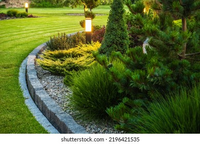 Landscaped Garden at the Backyard with Evenly Mowed Lawn and Island Bed with a Variety of Shrubs, Bushes, Trees and Evergreen Plants. Decorated with Bollard Outdoor Lighting Lamp.