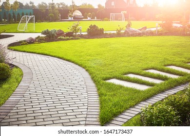 Landscaping of the garden. A tile path between green grass and a lawn with flowers in the sun. Soccer field in the background with copy space. - Shutterstock ID 1647173437
