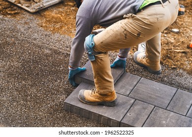 Landscaping Paver Worker Laying Paving Stones On Sandy Ground Of Construction Patio Yard Site In Spring Summer. Contractor Wearing Safety Protective Cloth, Gloves And Knee Pads For Installation Work. 