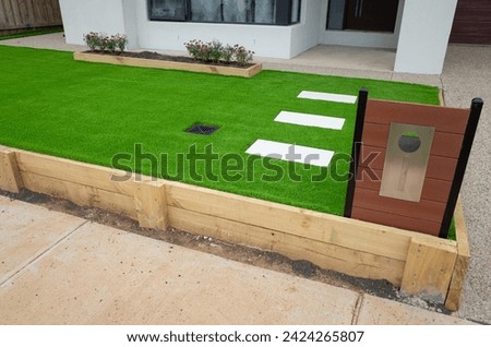 Landscaping project of artificial grass or synthetic turf with a wooden raised garden bed, stepping stones and a mailbox in a front yard outside an Australian residential suburban home. 