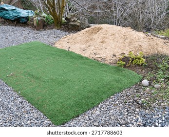 Landscaping project in progress featuring a rectangular patch of artificial green turf surrounded by gray gravel and a large pile of light brown sand in a backyard garden setting.