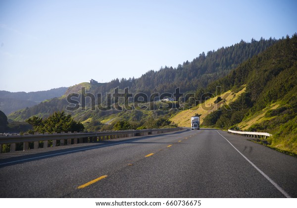 Landscaping\
with semi truck with the trailer is moving from the horizon on an\
empty highway among the green hills and\
fens