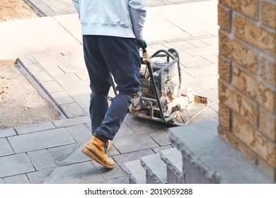 Landscaping Worker Contractor Using Plate Compactor To Apply Polymeric Joint Sands On Interlock Pavers. Home Garden And Landscape Renovation Project Background. 