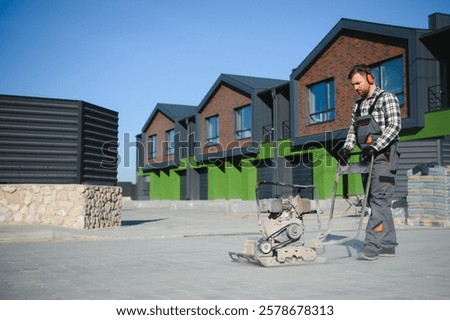 Landscaping worker using vibratory tamping machine plate compactor on a new patio driveway pavement to apply Polymeric sand to fill joints. Home garden and landscape renovation project background.